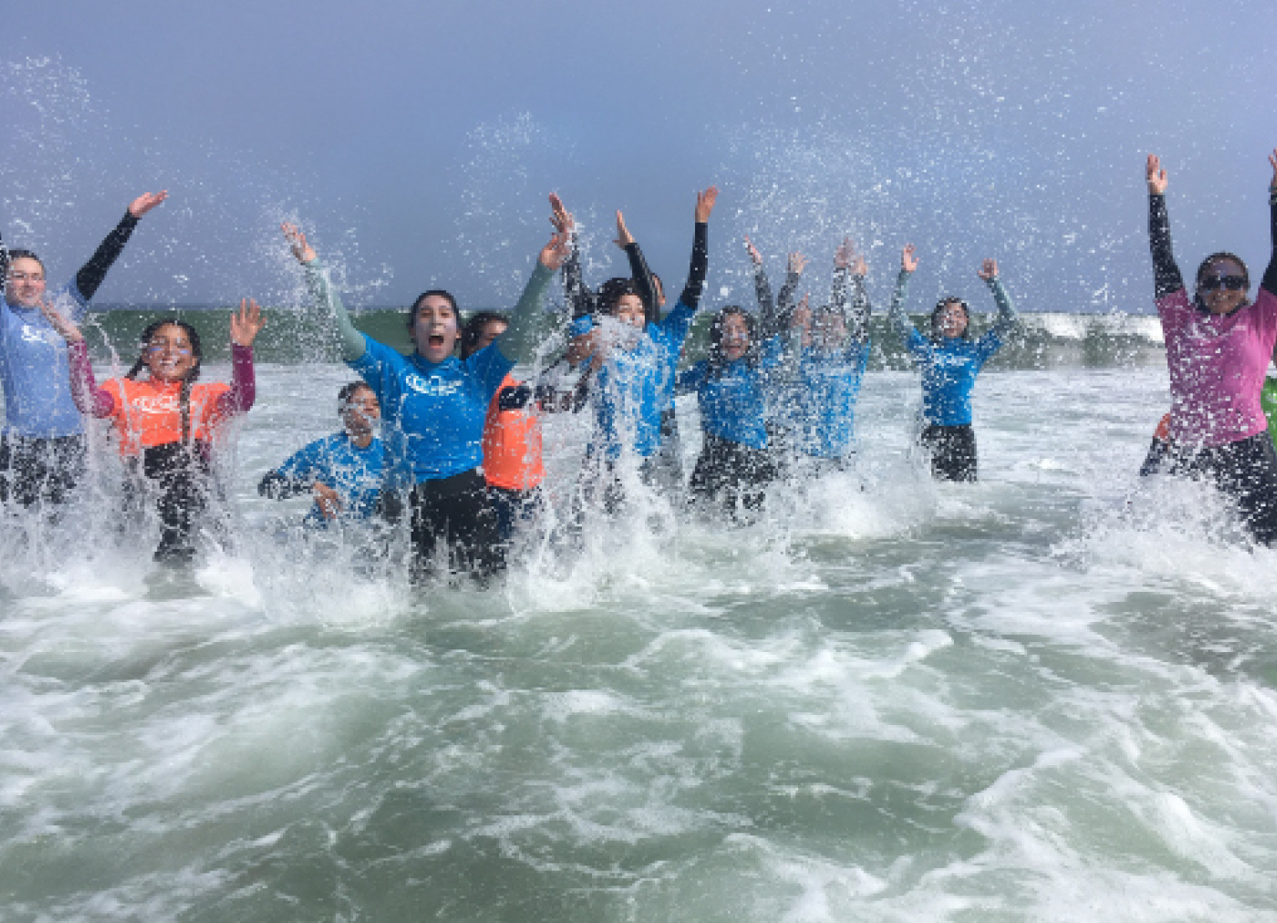 A group of teenagers in wetsuits jumping and raising their hands in the air in waist deep water