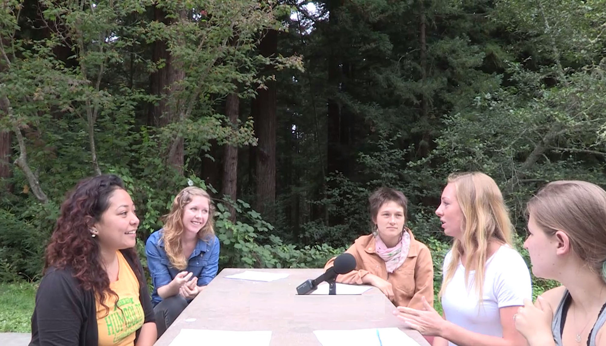 5 women sitting at a table outside speaking into a microphone with a forest in the background